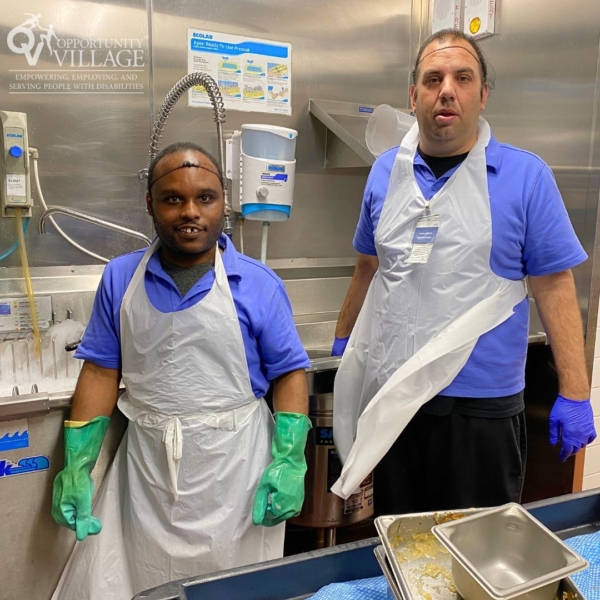 two workers in a commercial kitchen with aprons and gloves on.